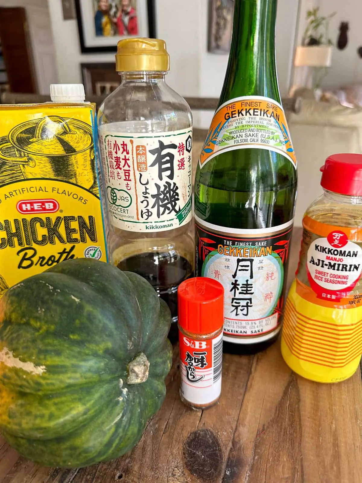 An acorn squash, carton of chicken broth, bottle of soy sauce, bottle of sake, mirin, and Ichimi Togarashi Japanese Chili Pepper on a wooden table.