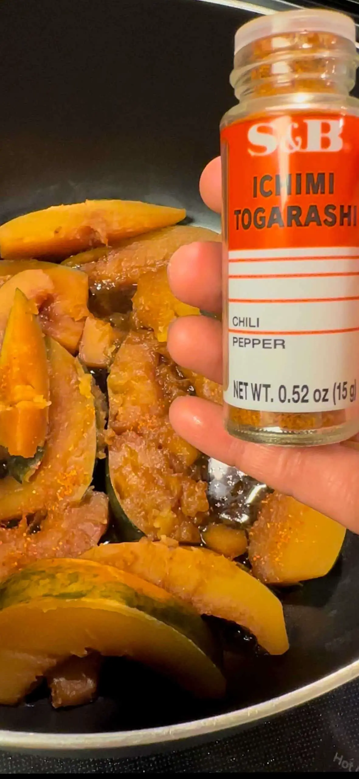 Cooked wedges of acorn squash in a soy braising liquid and a hand holding a bottle of Ichimi Togarashi in the foreground.