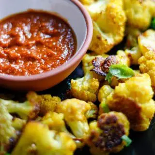 Tender Cauliflower florets roasted with Indian spices garnished with cilantro and a small terracotta bowl with red chutney.