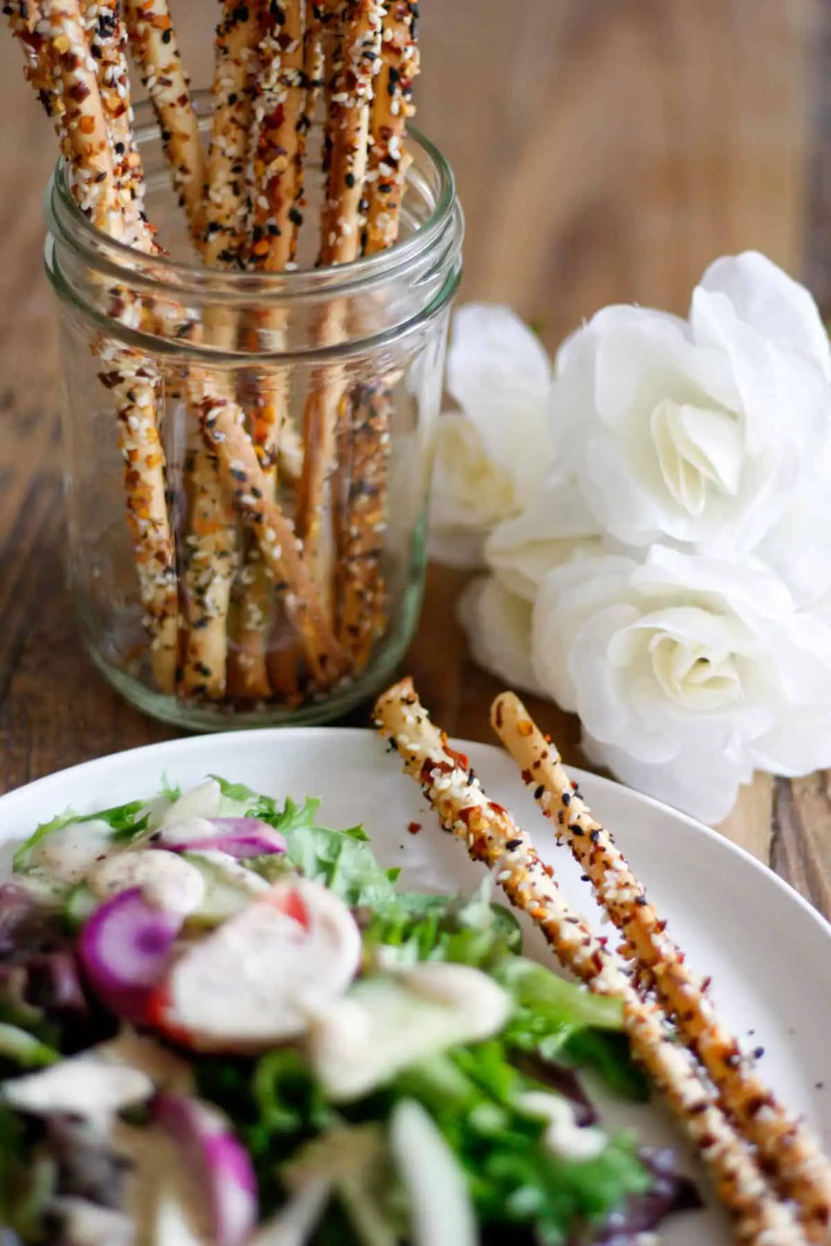 Thin breadsticks coated with everything bagel seasoning, salt and red chili flakes standing in a glass jar with white roses to the right of the jar, and a white plate with a green leaf salad and purple radishes and 2 breadsticks.
