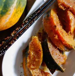 Wedges of cooked acorn squash with sprinkles of Japanese chili pepper on a white dish with chopsticks resting on the side, an acorn squash to the left of the white dish.