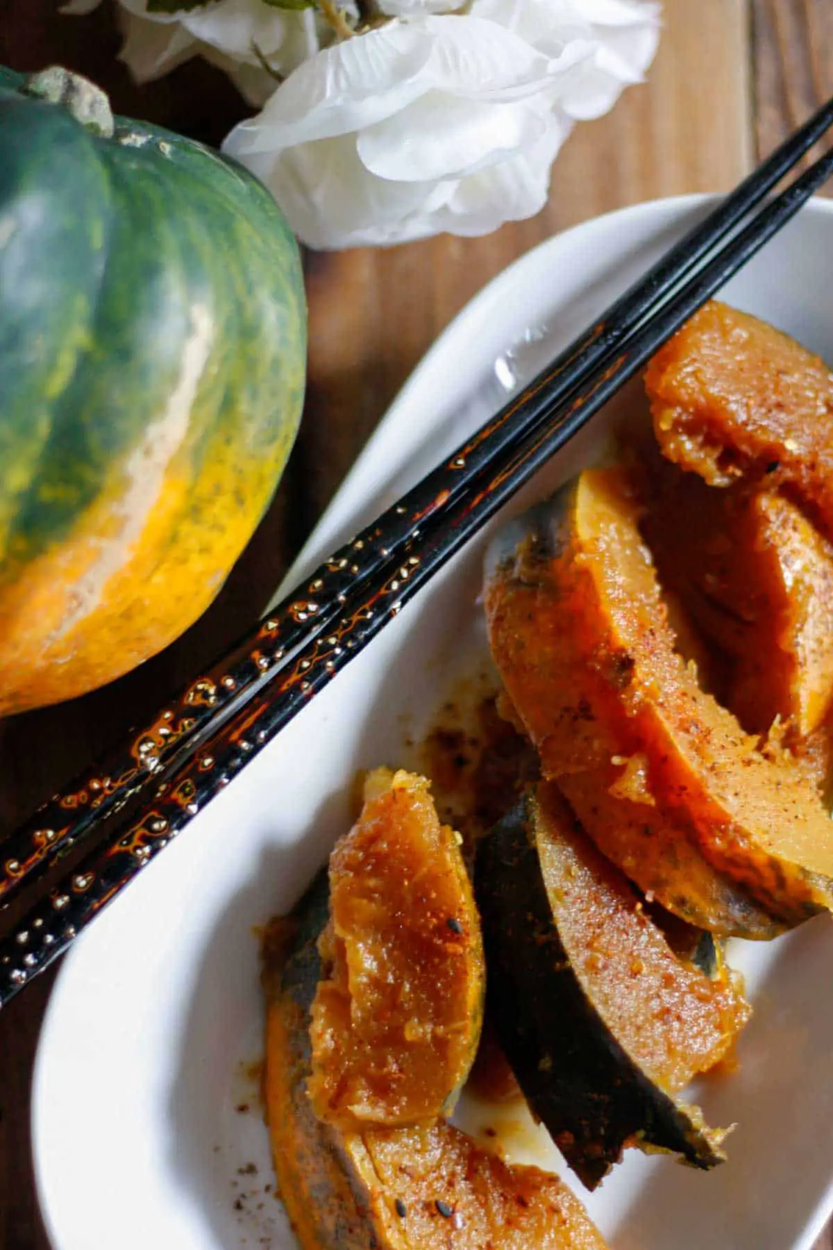 Wedges of cooked acorn squash with sprinkles of Japanese chili pepper on a white dish with chopsticks resting on the side, an acorn squash to the left of the white dish and a few white rose petals.