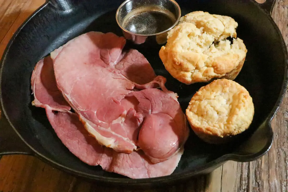 Slices of cooked country ham, silver bowl with red eye gravy, and 2 biscuits in a cast iron pan.