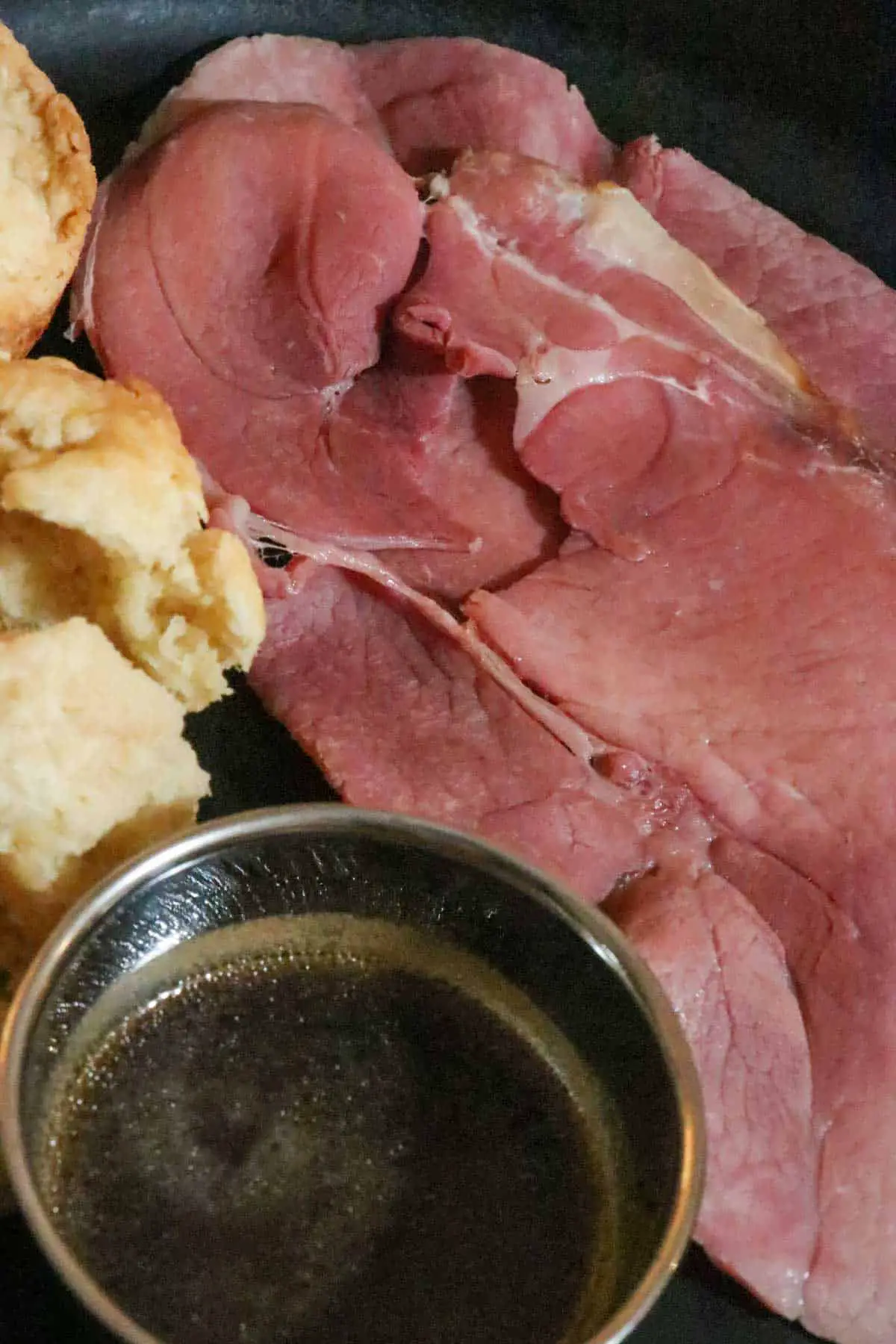 Cooked slices of country ham, a silver bowl with red eye gravy, and biscuits next to the ham.