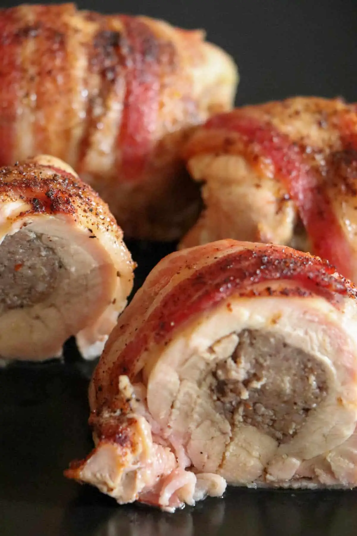 Chicken armadillo eggs on a black plate which is chicken wrapped with bacon and stuffed with sausage and one of the eggs has been sliced in two so you can see the 3 layers of sausage, chicken, and bacon.