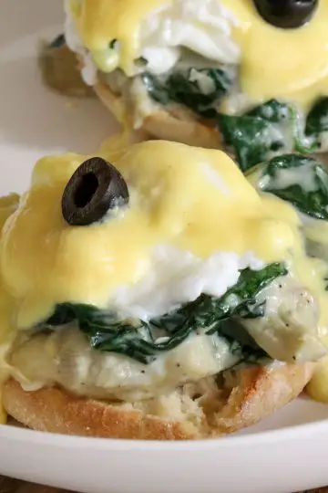 Poached eggs with hollandaise sauce and a black olive atop creamed spinach and artichokes and an English muffin on a white dish.