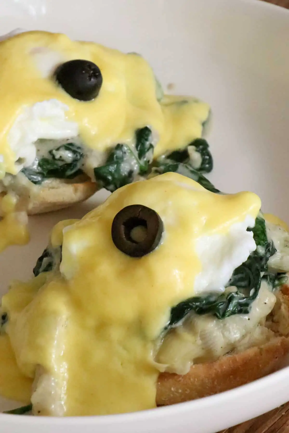 Poached eggs with hollandaise sauce and a black olive atop creamed spinach and artichokes and an English muffin on a white dish.