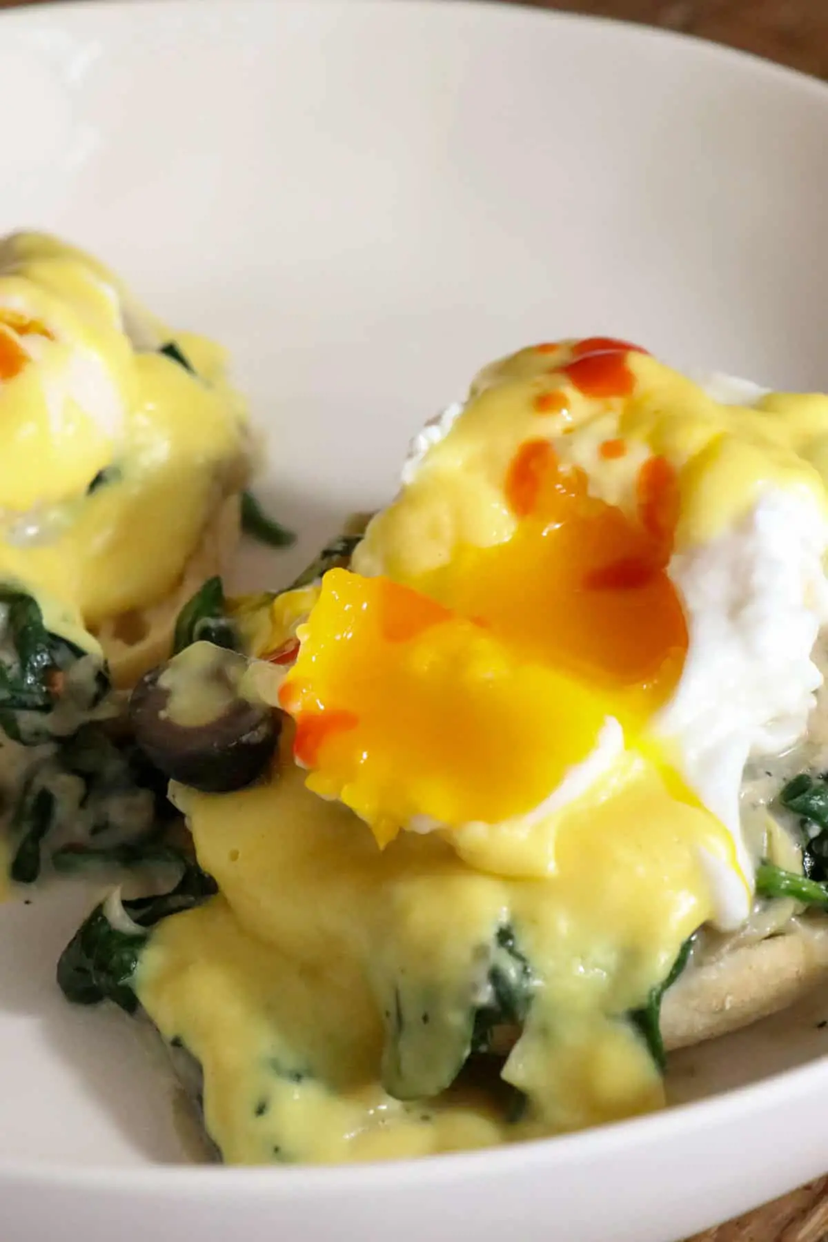 Poached eggs with hollandaise sauce and splashes of Tabasco and a black olive atop creamed spinach and artichokes and an English muffin on a white dish.
