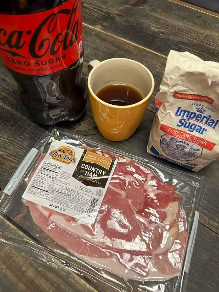 Country ham slices in a package, a bag of Imperial Sugar, a yellow coffee mug with black coffee, and a bottle of Coca Cola Zero Sugar.