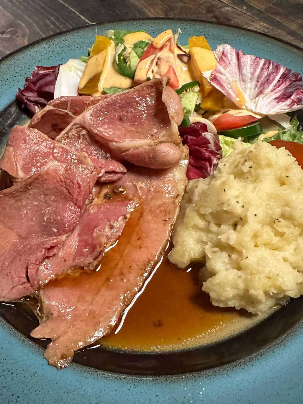 A blue rimmed plate containing country ham slices with red eye gravy poured over them served with mashed potatoes and a salad.