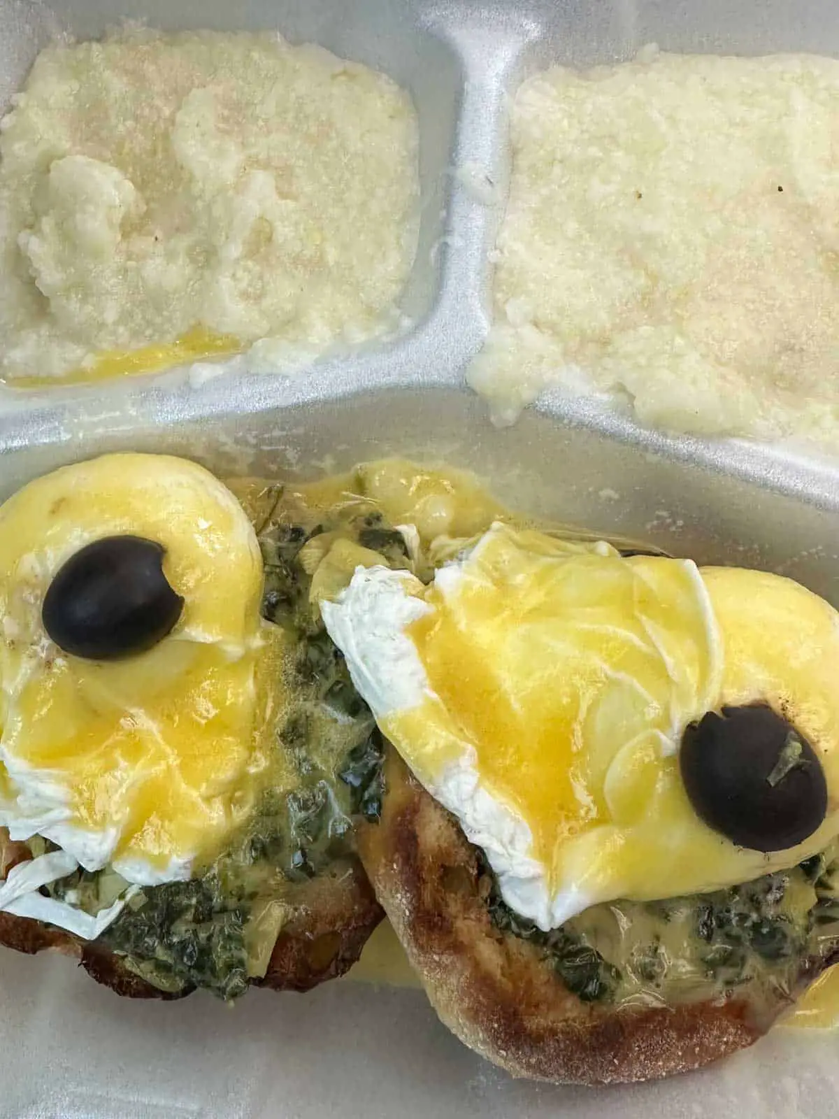 2 each Eggs Sardou which is poached egg on top of creamed spinach, artichoke, and English muffin and topped with Hollandaise sauce and a black olive, with grits in the background.