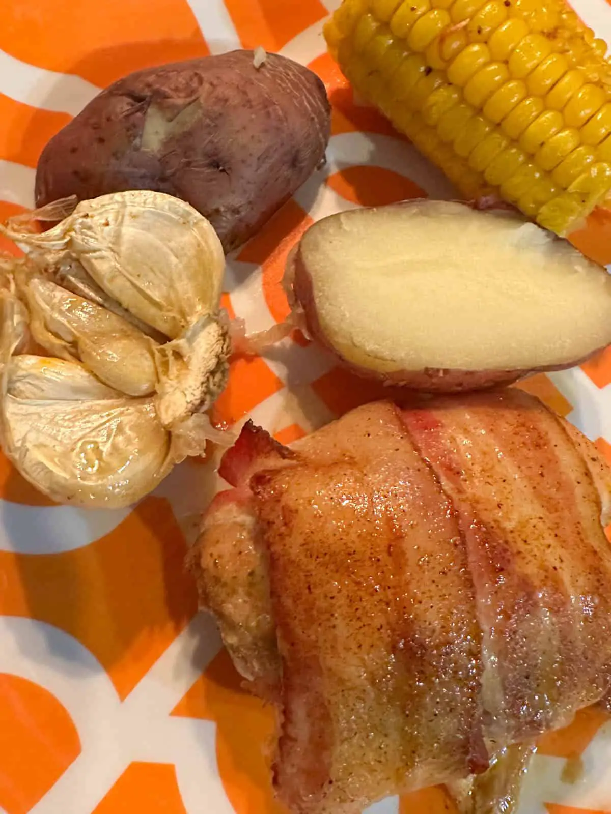 Chicken armadillo egg, half a bulb of garlic, halves of a red potato and some corn on an orange and white plate.