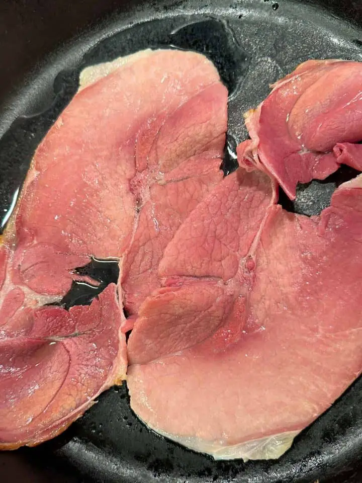 2 slices of country ham cooking in a cast iron skillet.