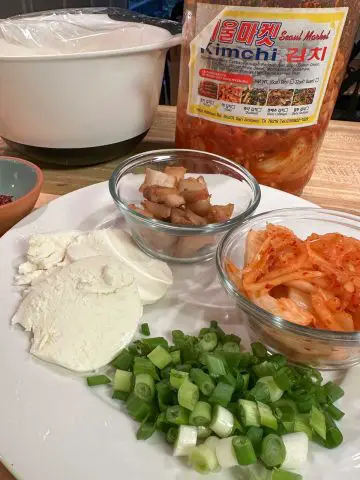White mixing bowl covered with plastic wrap, a large jar of kimchi, the edge of a blue bowl with gochujang, a white plate topped with slices of mozzarella, diced kimchi, pork belly, and green onions.