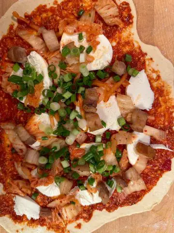 Uncooked pizza base topped with slices of mozzarella, diced cooked pork belly, diced kimchi and green onions, and a tomato sauce.