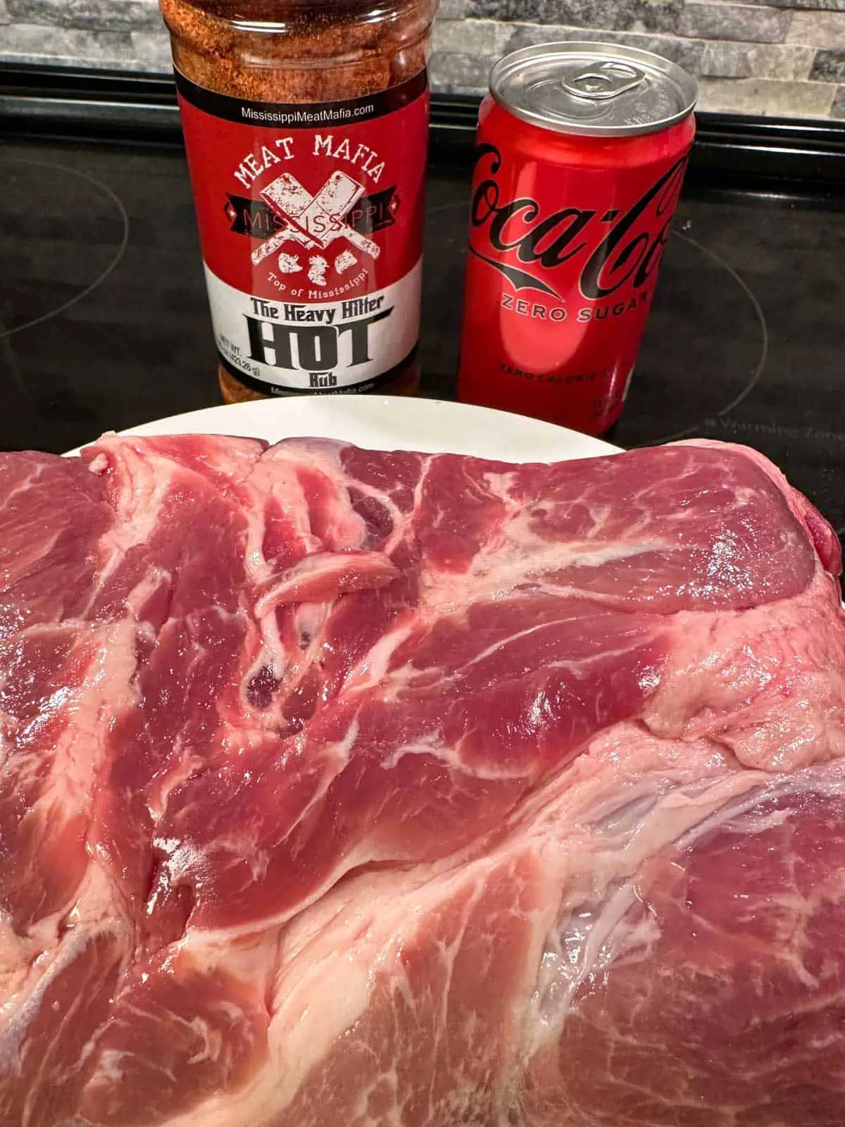 A large piece of pork butt on a white plate, a bottle of Meat Mafia The Heavy Hitter Hot Rub, and a can of Coca Cola Zero.