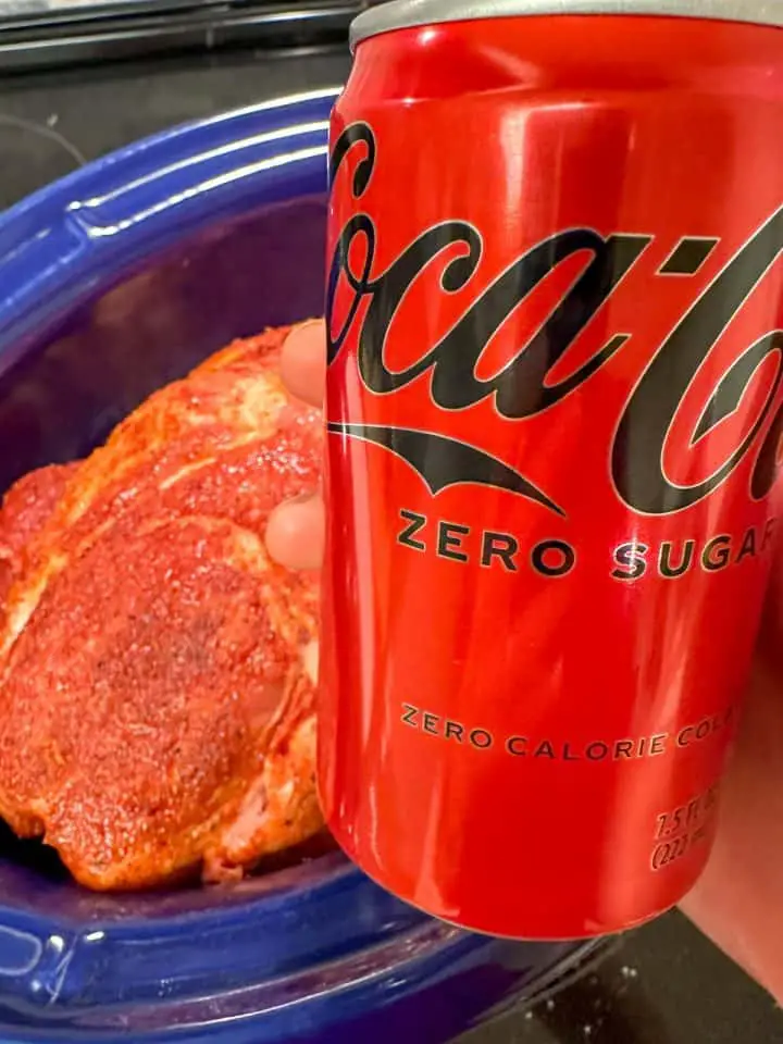Seasoned uncooked pork butt in a blue slow cooker and a can of Coca Cola Zero in the foreground.