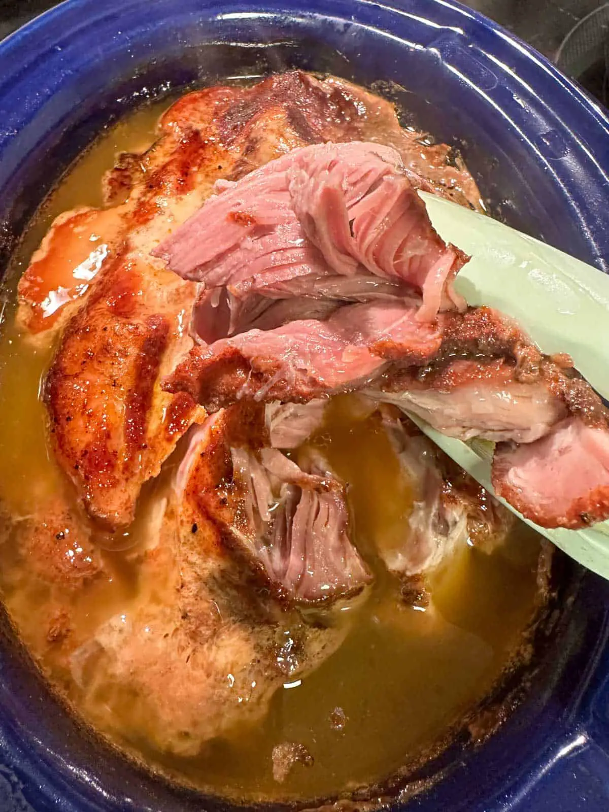 Tender pulled pork in a blue slow cooker with broth from cooking and a pair of blue tongs holding some of the cooked pulled pork.