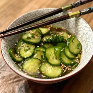 Pickled slices of cucumber garnished with sesame seeds and green onions in a small patterned bowl with chopsticks resting on the bowl and the bowl is on a wooden board.