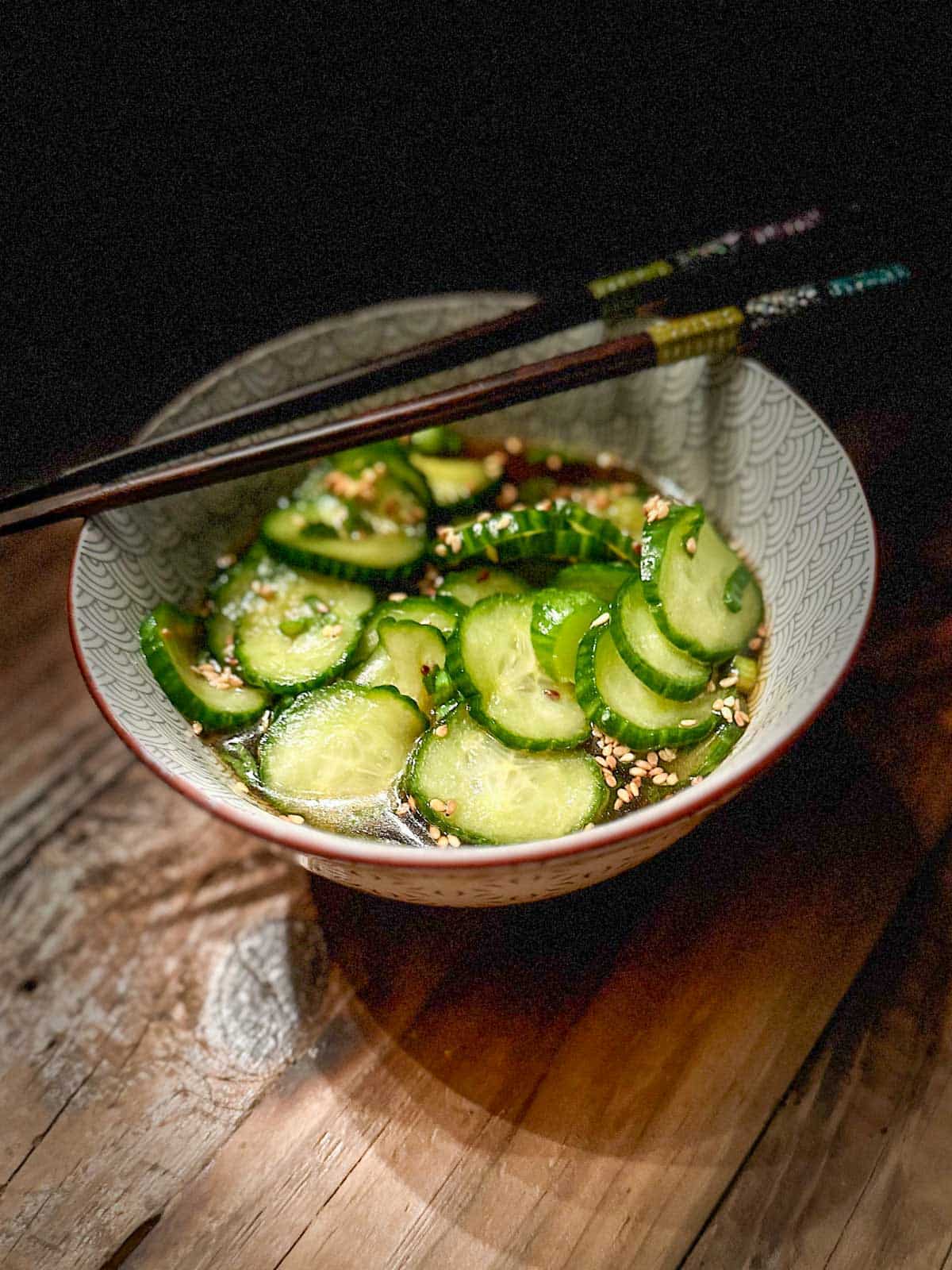 Pickled slices of cucumber garnished with sesame seeds and green onions in a small patterned bowl with chopsticks resting on the bowl and the bowl is on a wooden board.