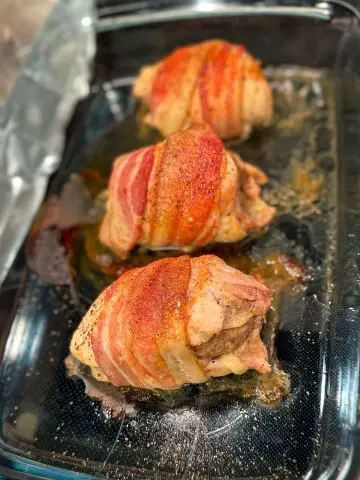 Three chicken armadillo eggs which are chicken thighs stuffed with sausage, wrapped with bacon and sprinkled with dry rub on a glass dish with the fats from cooking.