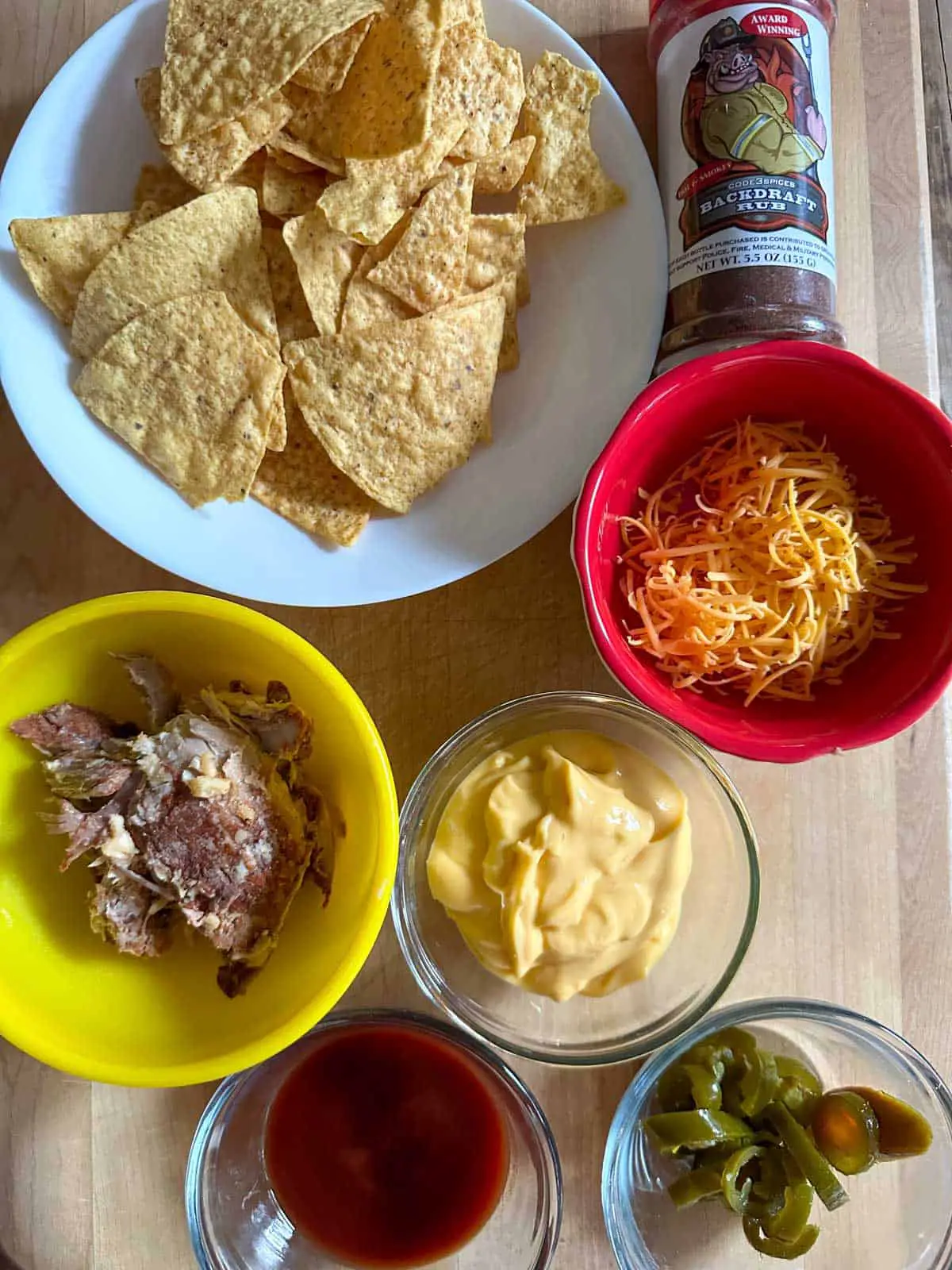 Tortilla chips in a white bowl, a bottle of Backdraft Rub, shredded cheese in a red bowl, pulled pork in a yellow bowl, and nacho cheese sauce, barbecue sauce and jalapeños in small glass bowls.