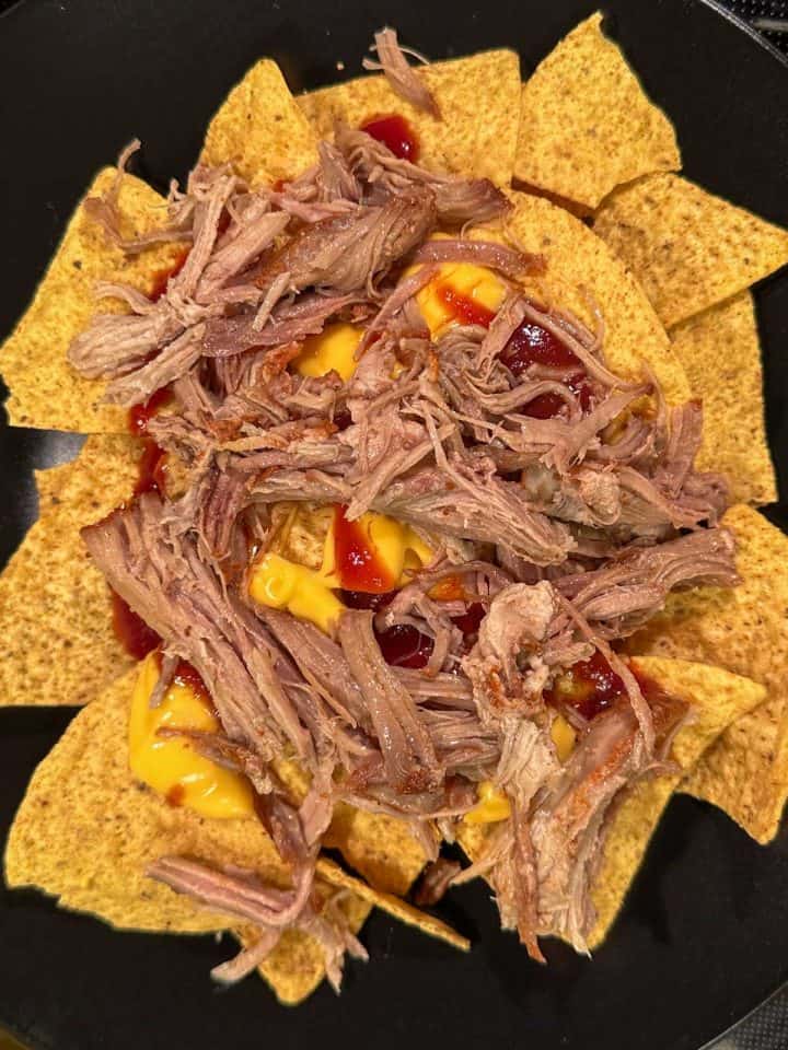 Tortilla chips topped with cheese sauce, pulled pork, and barbecue sauce on a black plate.