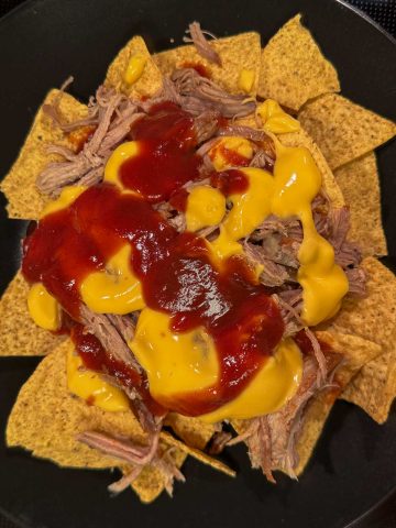Tortilla chips topped with cheese sauce, pulled pork, and barbecue sauce on a black plate.