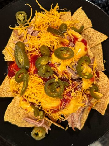 Tortilla chips topped with cheese sauce, pulled pork, shredded cheese, jalapeños and barbecue sauce on a black plate.