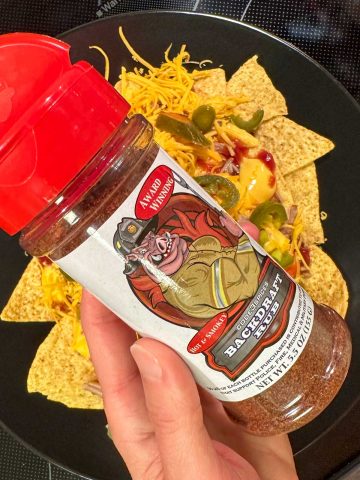 Tortilla chips topped with cheese sauce, shredded cheese, barbecue sauce, jalapenos and pulled pork on a black plate with a bottle of dry rub called Backdraft Rub being held by a hand in the foreground.