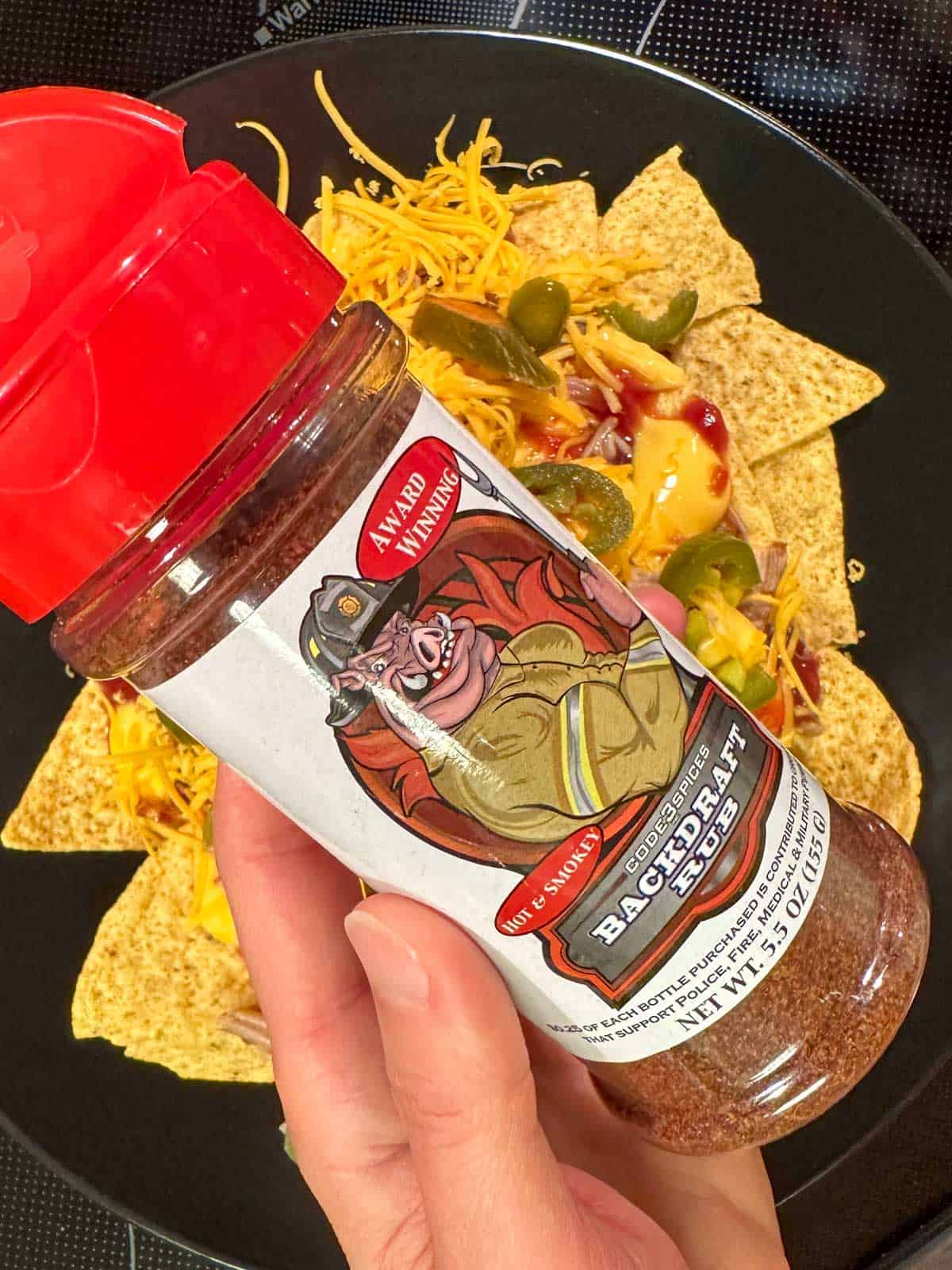 Tortilla chips topped with cheese sauce, shredded cheese, barbecue sauce, jalapenos and pulled pork on a black plate with a bottle of dry rub called Backdraft Rub being held by a hand in the foreground.