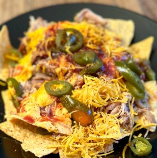 Tortilla chips topped with cheese sauce, shredded cheese, barbecue sauce, jalapeños and pulled pork on a black plate with dry rub sprinkled on top.