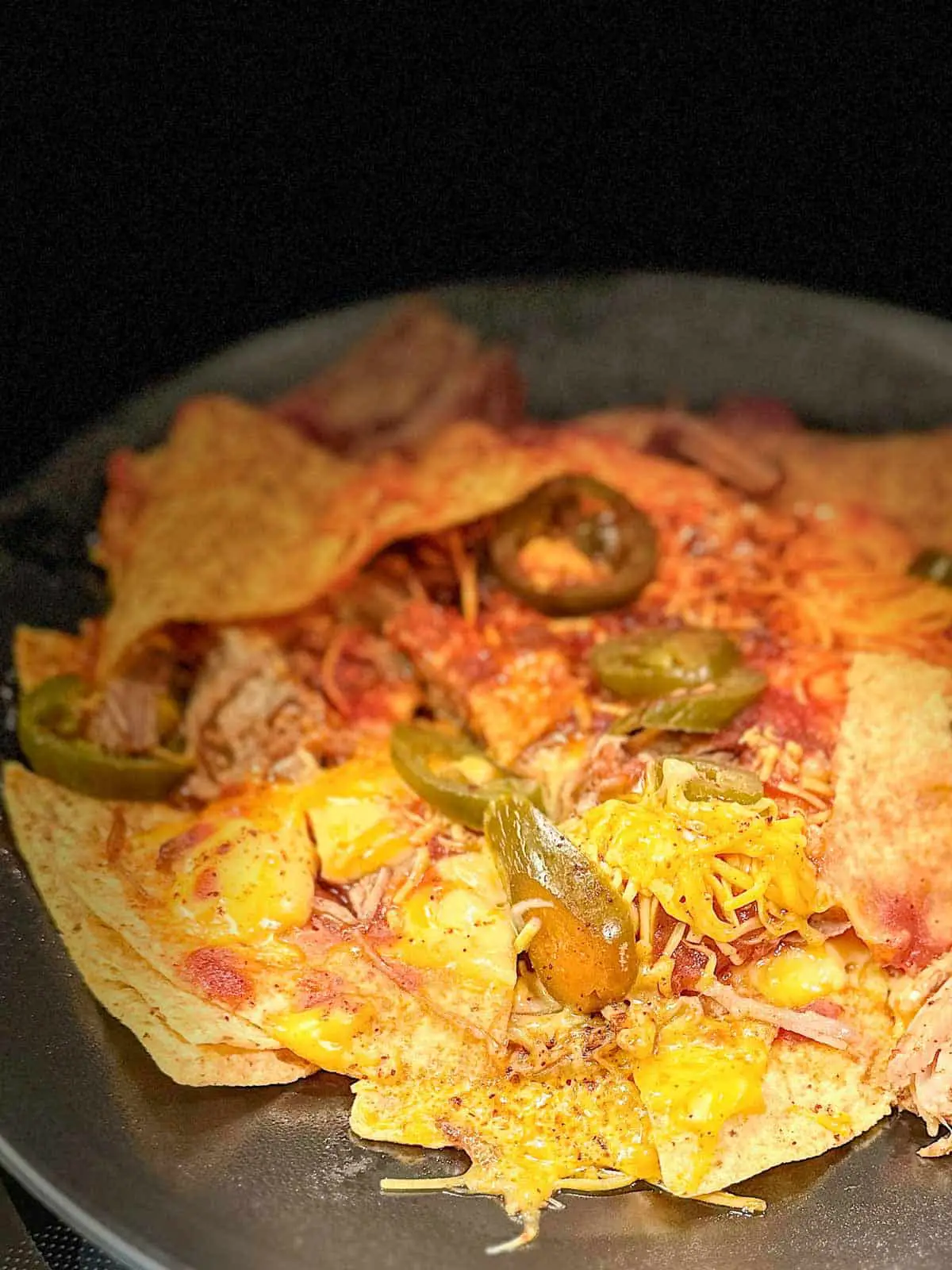 Tortilla chips topped with cheese sauce, shredded cheese, barbecue sauce, jalapeños and pulled pork on a black plate with dry rub sprinkled on top.