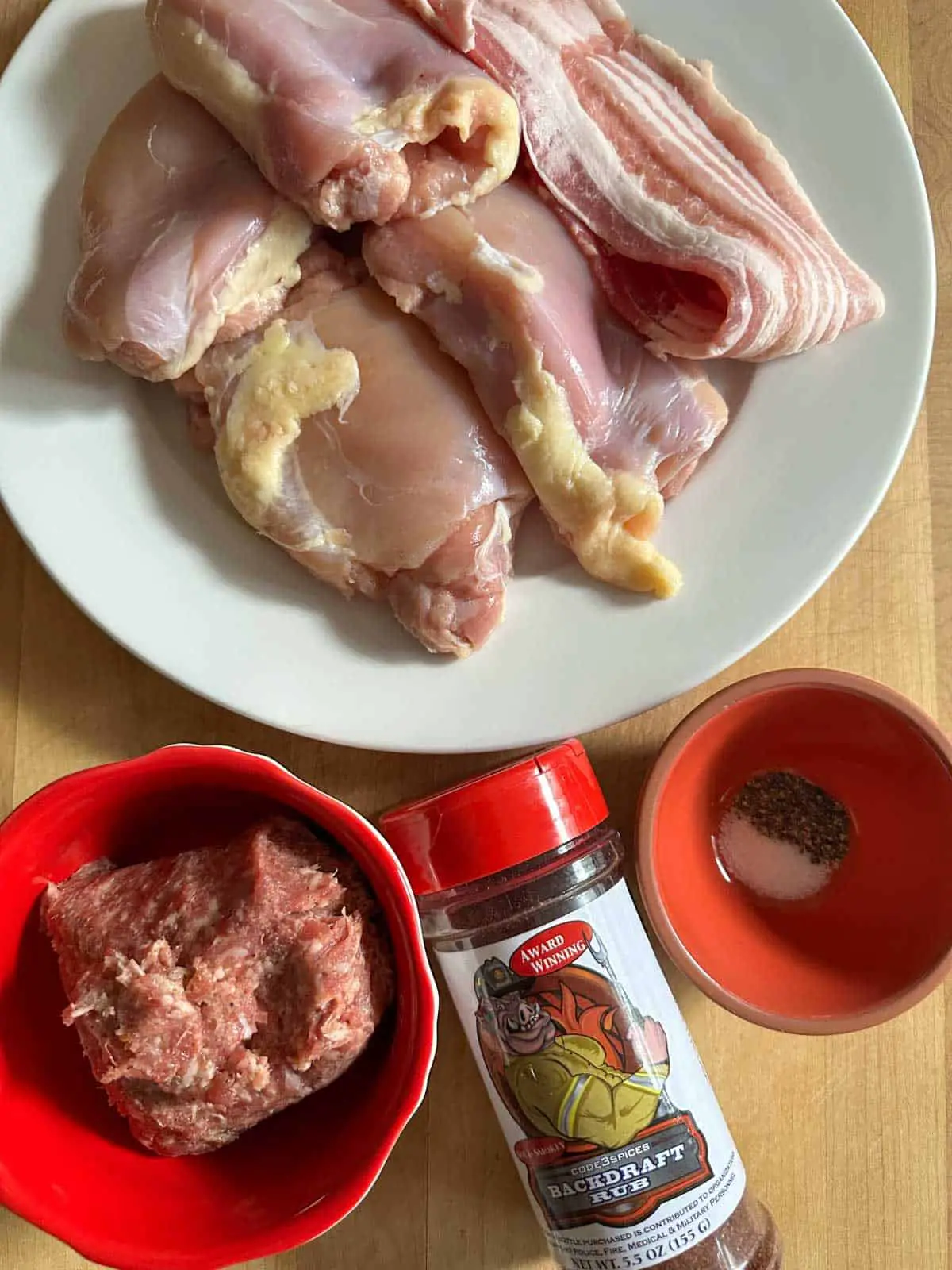 Chicken thighs and pieces of bacon on a white plate, breakfast sausage in a red bowl, a small bowl with salt and pepper, and a bottle of Backdraft Rub.
