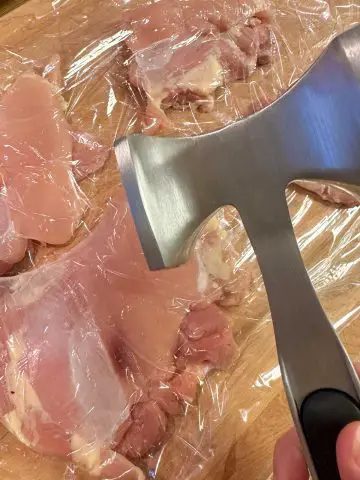 Chicken thighs laying between plastic wrap with a meat mallet poised over the chicken thighs.