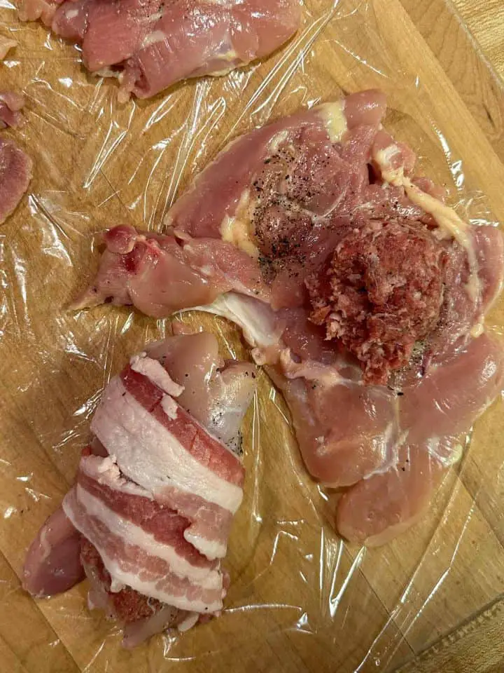 Uncooked pieces of chicken thighs on plastic wrap one is laying flat with sausage on top, and one has been rolled and wrapped with bacon.