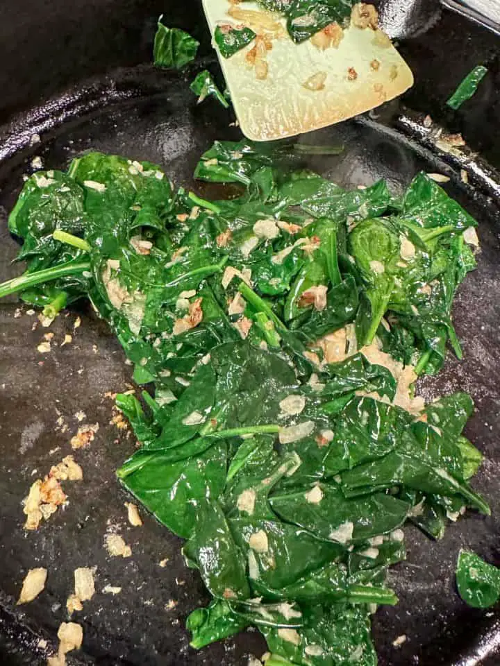 Wilted baby spinach and sautéed garlic and onion in a cast iron pan with a blue silicone utensil resting on the pan.