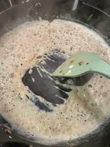 Flour, butter, and milk combining to make a roux in a cast iron pan with a blue silicone utensil.