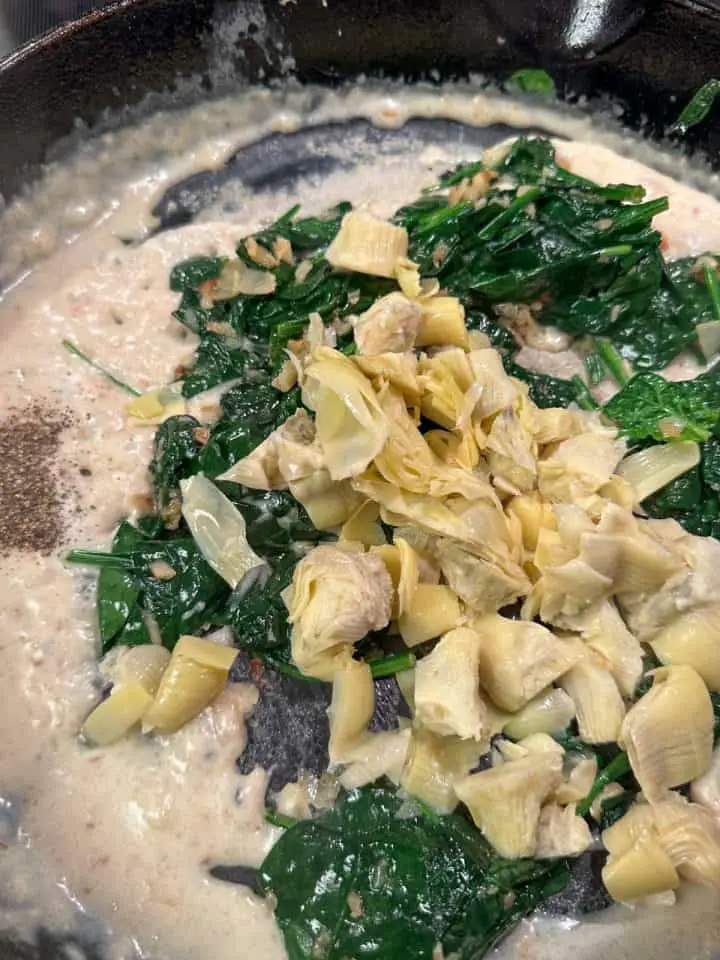 A cast iron pan containing a white roux, chopped artichoke hearts, wilted spinach, and salt and pepper.