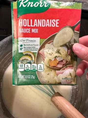 A packet of Knorr Hollandaise Sauce Mix poised over a pan with the prepared Hollandaise sauce and a blue whisk.