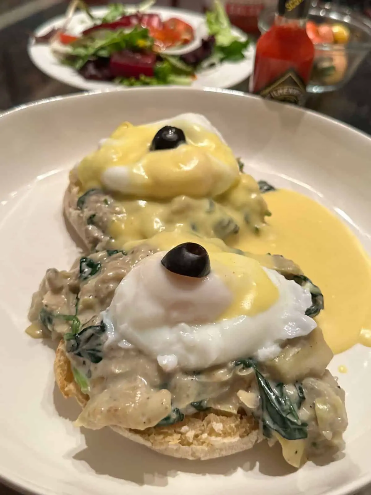 Poached eggs with hollandaise sauce and black olives atop creamed spinach and artichokes and an English muffin on a white dish with a salad, fruit, and bottle of Tabasco in the background.