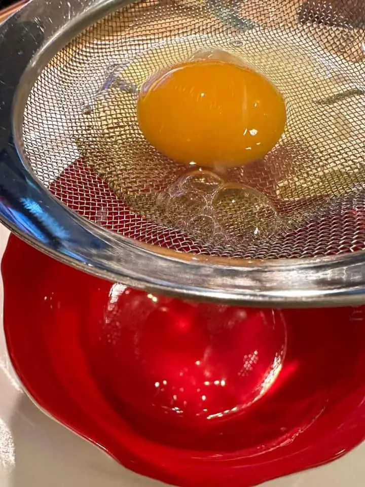 A mesh strainer containing an uncooked egg poised over a red bowl.