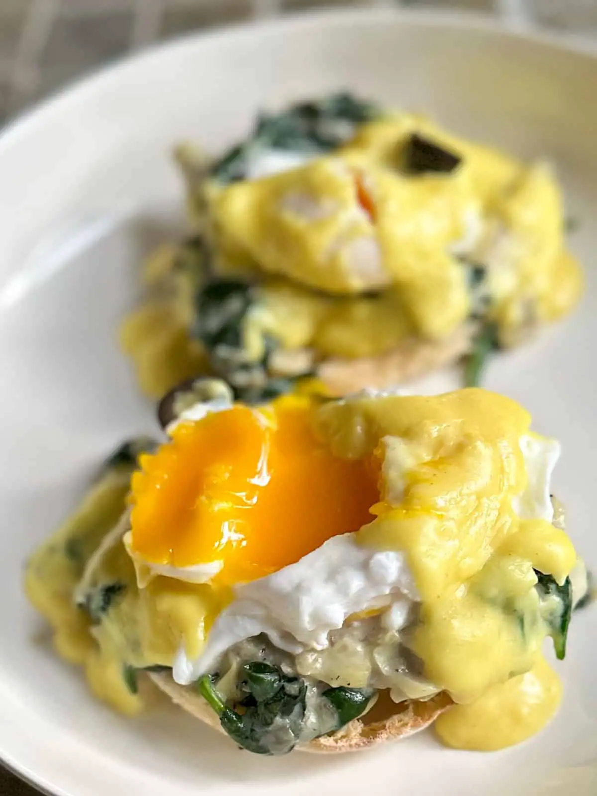 Poached eggs with hollandaise sauce and a black olive atop creamed spinach and artichokes and an English muffin on a white dish with one of the poached eggs showing the egg yolk.