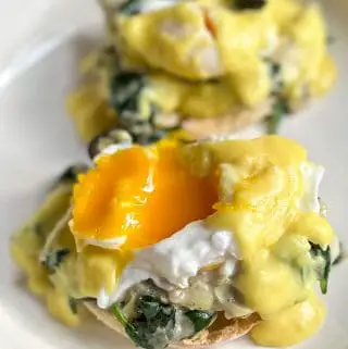 Poached eggs with hollandaise sauce and a black olive atop creamed spinach and artichokes and an English muffin on a white dish with one of the poached eggs showing the egg yolk.