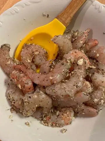 Uncooked shrimp tossed with minced garlic and seasonings in a white bowl with a yellow wooden handled spoon resting in the bowl.