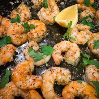 Pieces of cooked shrimp garnished with parsley and wedges of lemon in a cast iron pan.