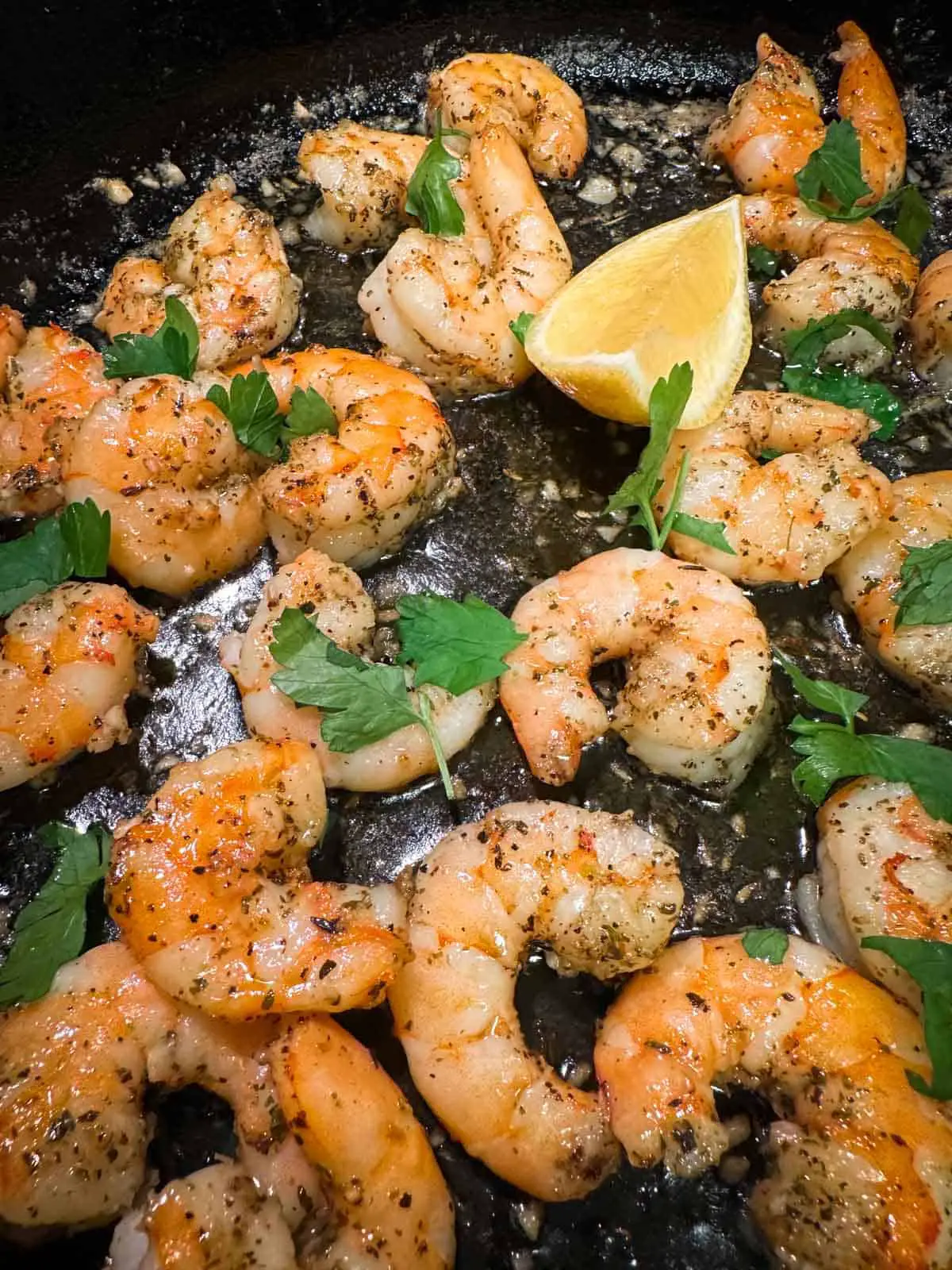 Pieces of cooked shrimp garnished with parsley and wedges of lemon in a cast iron pan.