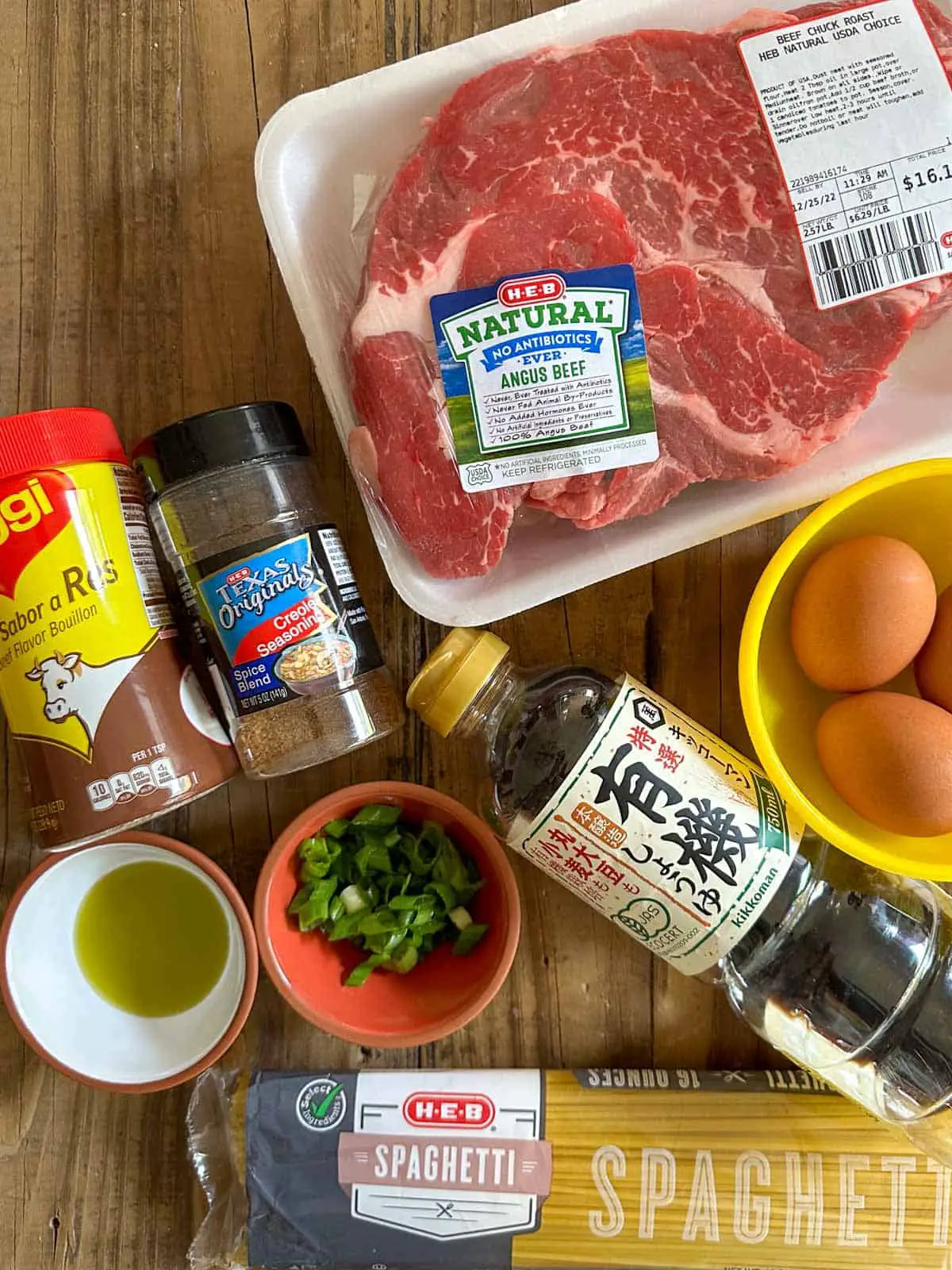 A package of beef chuck roast, Maggi beef bouillon seasoning, Creole seasoning, olive oil in a small bowl, minced green onions in a small bowl, bottle of soy sauce, package of spaghetti, and a yellow bowl containing eggs.