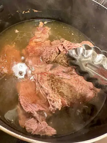 A pot containing cooked beef chuck roast and beef broth with a pair of tongs holding some of the tender beef.