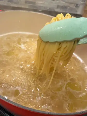 A pair of tongs holding some cooked spaghetti over a pot filled with water and cooked spaghetti.